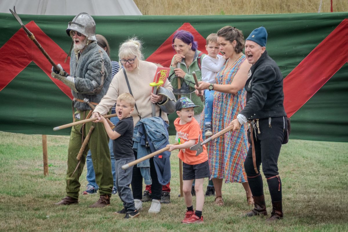 A group of historical re-enactors and excited families, including children, participating in a ground combat or training demonstration with wooden spears/pikes, standing in front of the red and green barrier.