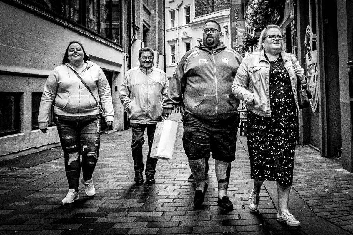 Black and white photo of four people walking together toward the camera down a narrow, brick-paved street with historic buildings.