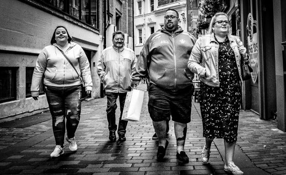Black and white photo of four people walking together toward the camera down a narrow, brick-paved street with historic buildings.