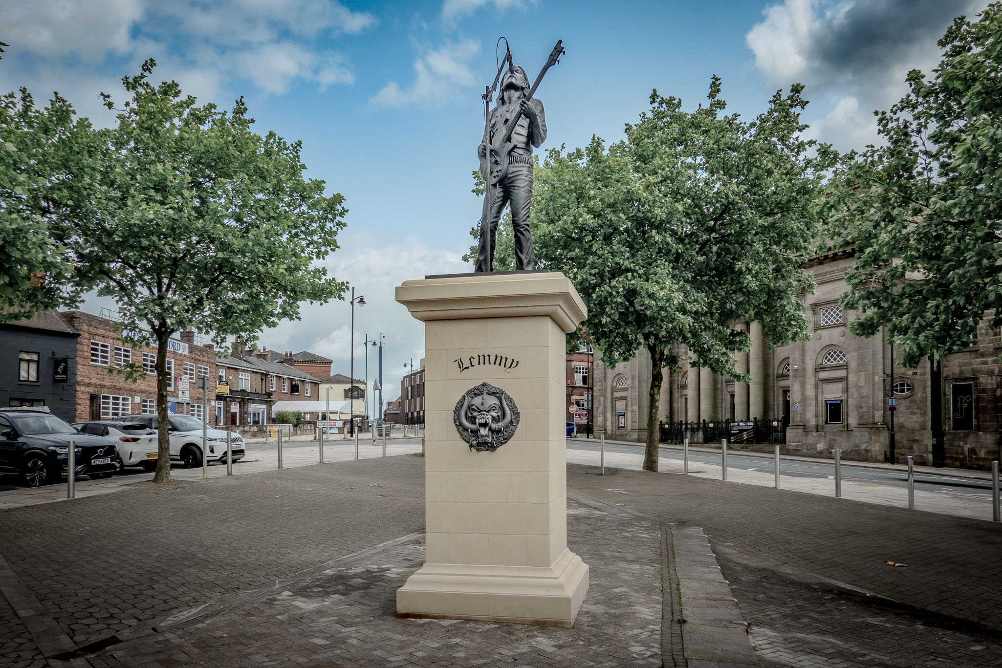 Full view of the Lemmy Kilmister bronze statue on a stone plinth in Burslem's market square under a partly cloudy sky.