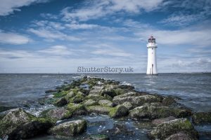 A white stone lighthouse standing in the sea at the end of a moss-covered rocky breakwater under a blue sky.