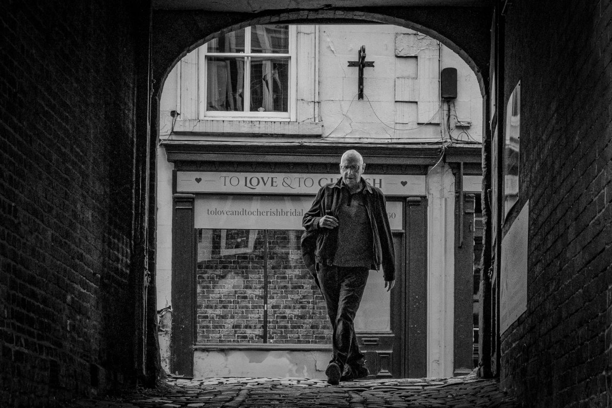 Black and white photo of an older man walking into a dark, brick archway onto a cobbled street with a shop window that says "TO LOVE & TO CHERISH" in the background in Ashbourne.