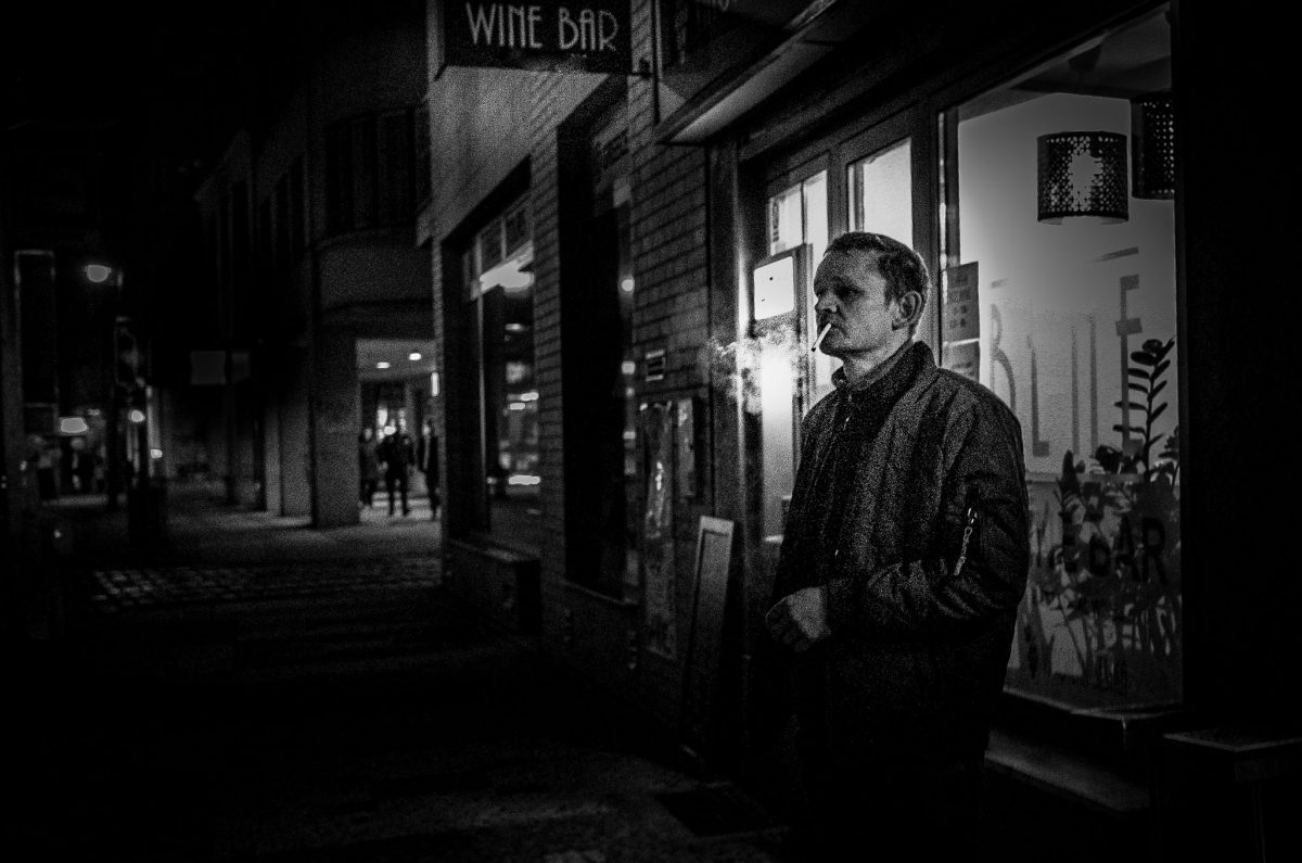 High-contrast black and white photo of a man smoking a cigarette outside a wine bar window at night, with smoke visible around his head.