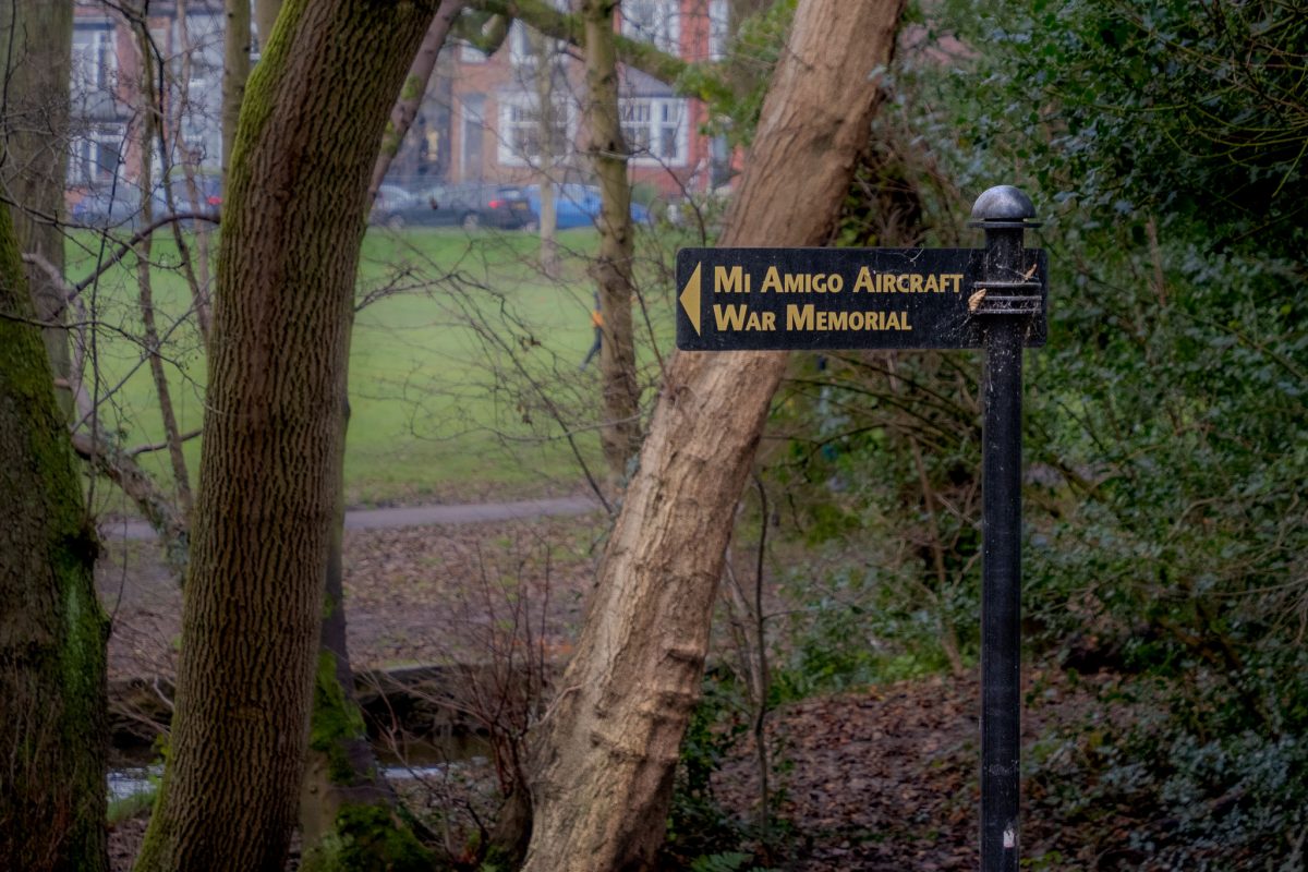 A black sign with gold lettering that reads 'Mi Amigo Aircraft War Memorial' standing among trees in a park.