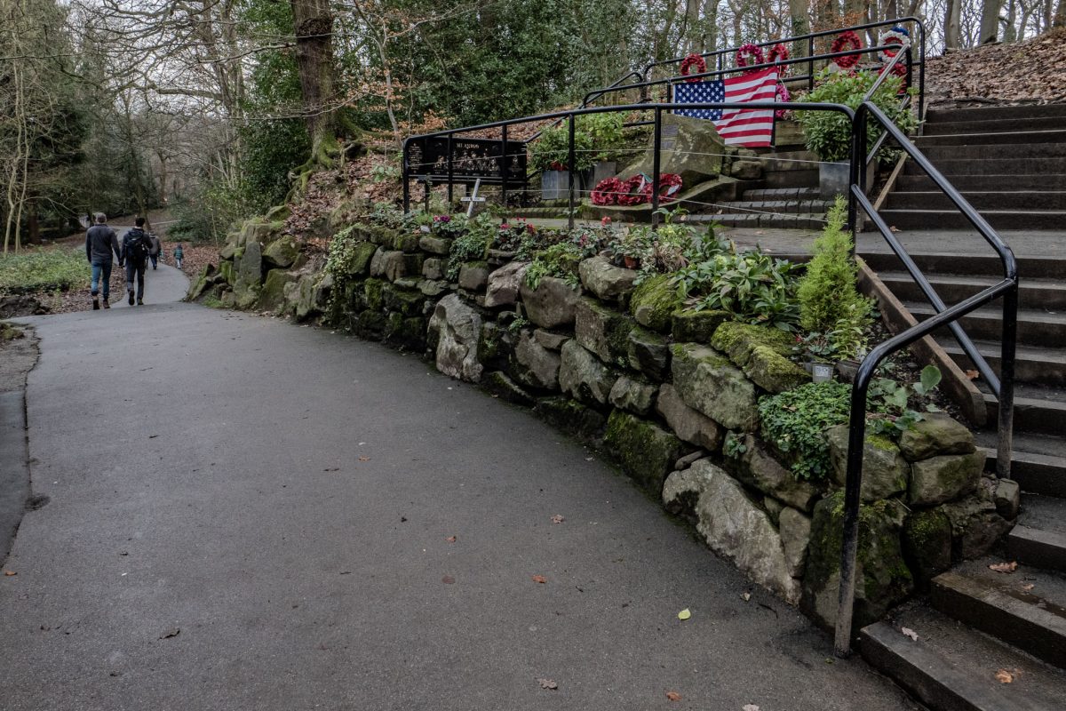 A paved path in a park leading past a stone wall and a memorial site decorated with American flags and poppy wreaths.
