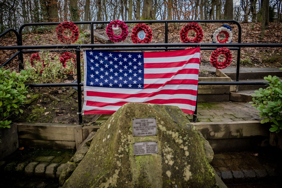 A large stone with bronze plaques sits in front of an American flag and several red poppy wreaths in a wooded area.