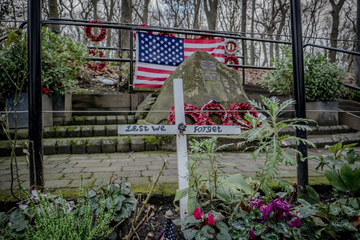 A white wooden cross with the words 'Lest We Forget' stands in a flower bed in front of the Mi Amigo memorial stone.