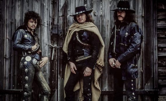Motorhead band members dressed in dark Western outlaw clothing, wearing wide-brimmed hats and bullet belts, standing in front of an old wooden wall.