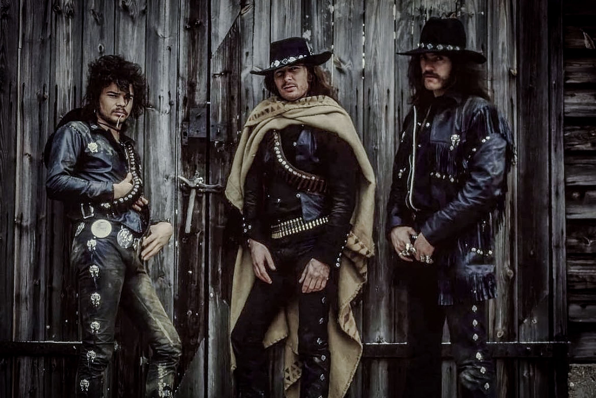 Motorhead band members dressed in dark Western outlaw clothing, wearing wide-brimmed hats and bullet belts, standing in front of an old wooden wall.