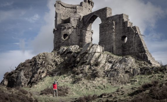Close-up of the crumbling stone walls, circular tower section, and high arch of the Mow Cop Castle folly, with a person taking a photo in the foreground.