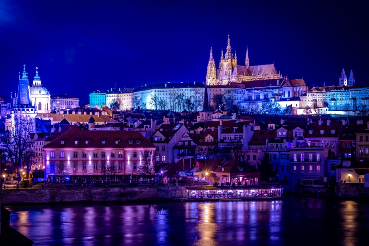 Panoramic night view of the Prague Castle complex and St. Vitus Cathedral illuminated in blue and purple light, overlooking the Vltava River and historic buildings.