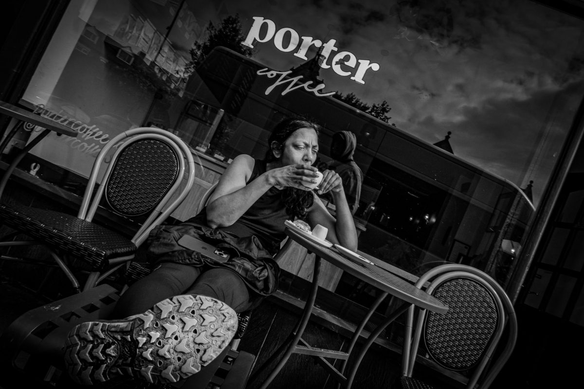 High-contrast black and white photo of a woman sitting alone, drinking coffee at an outdoor table with a view of the street reflected in the glass behind her, with the 'Porter Coffee' logo visible.
