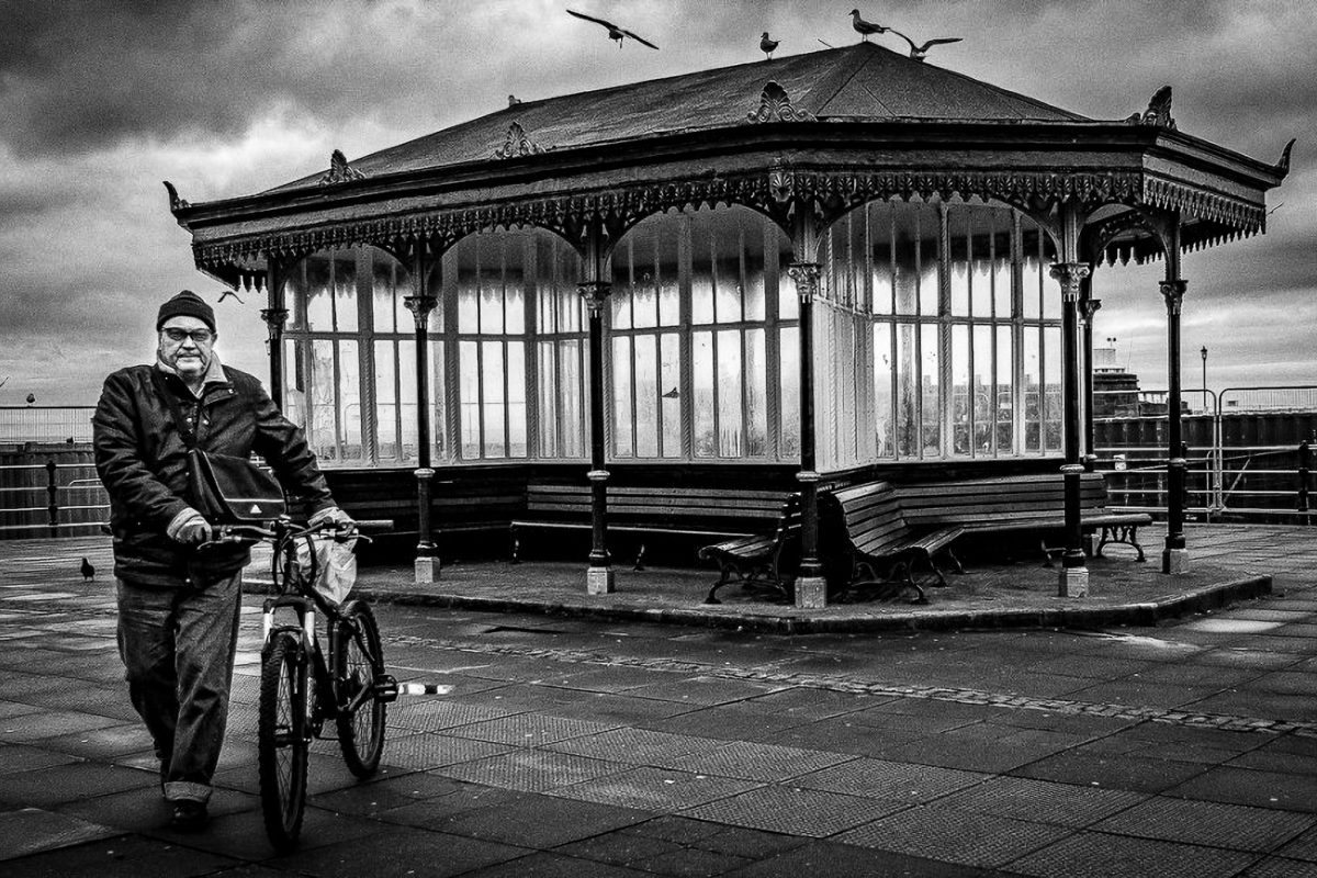 Black and white photo of a man walking a bicycle on the promenade past the historic Victorian shelter in New Brighton, Wirral. Seagulls fly above the roof.