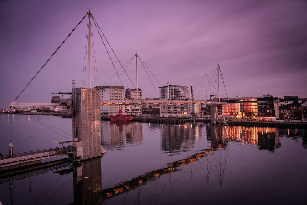 Royal Victoria Dock Bridge, a modern, cable-stayed footbridge in London, reflected in the calm waters of the dock at twilight with surrounding buildings illuminated.