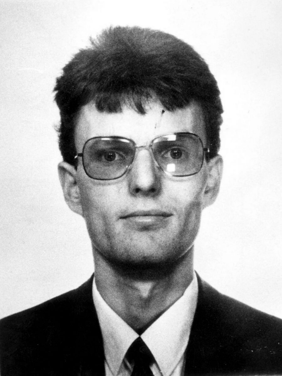 A black and white portrait of a young man with dark hair wearing large glasses and a suit with a tie.