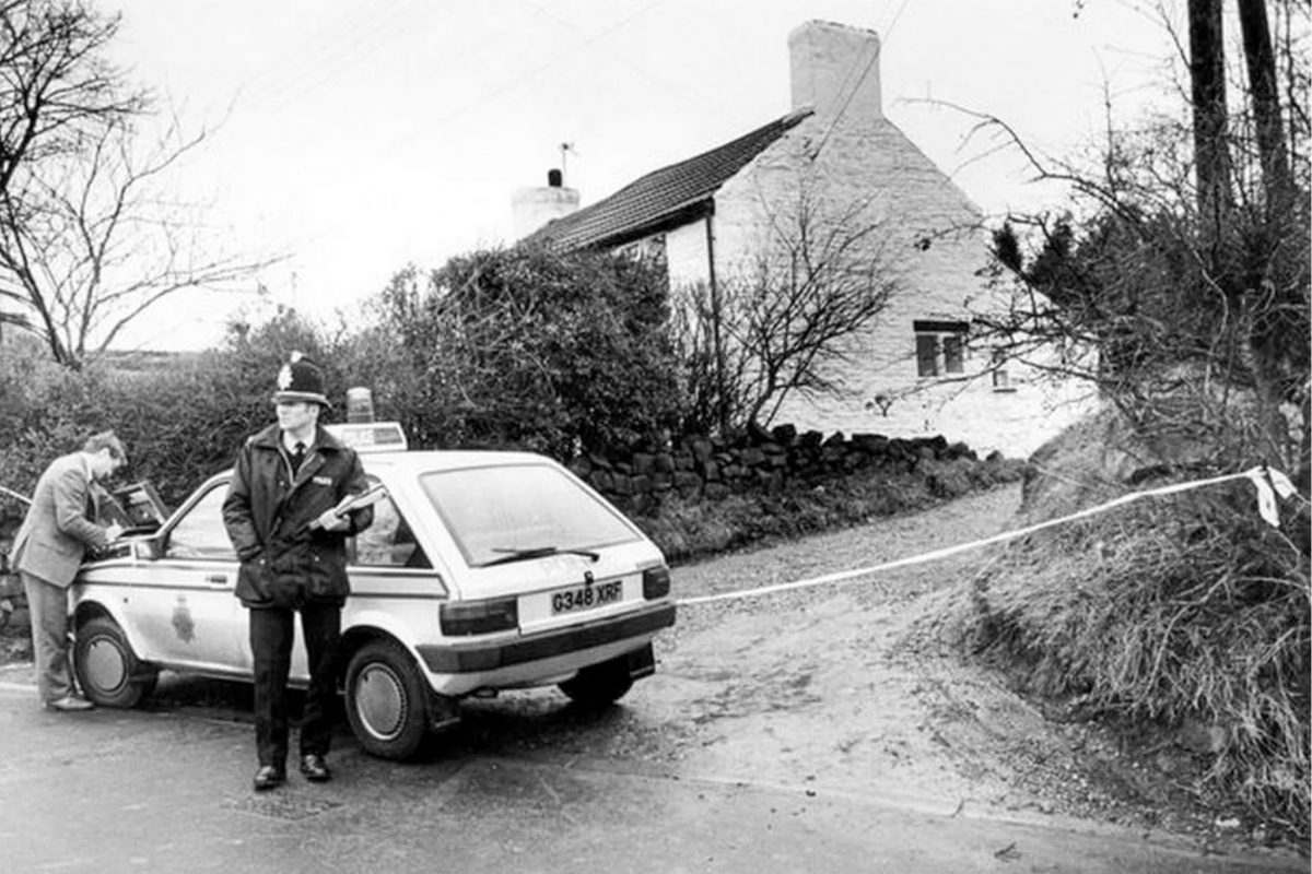 A vintage police officer standing next to a white police car with a cordon across a rural lane in front of a white cottage.