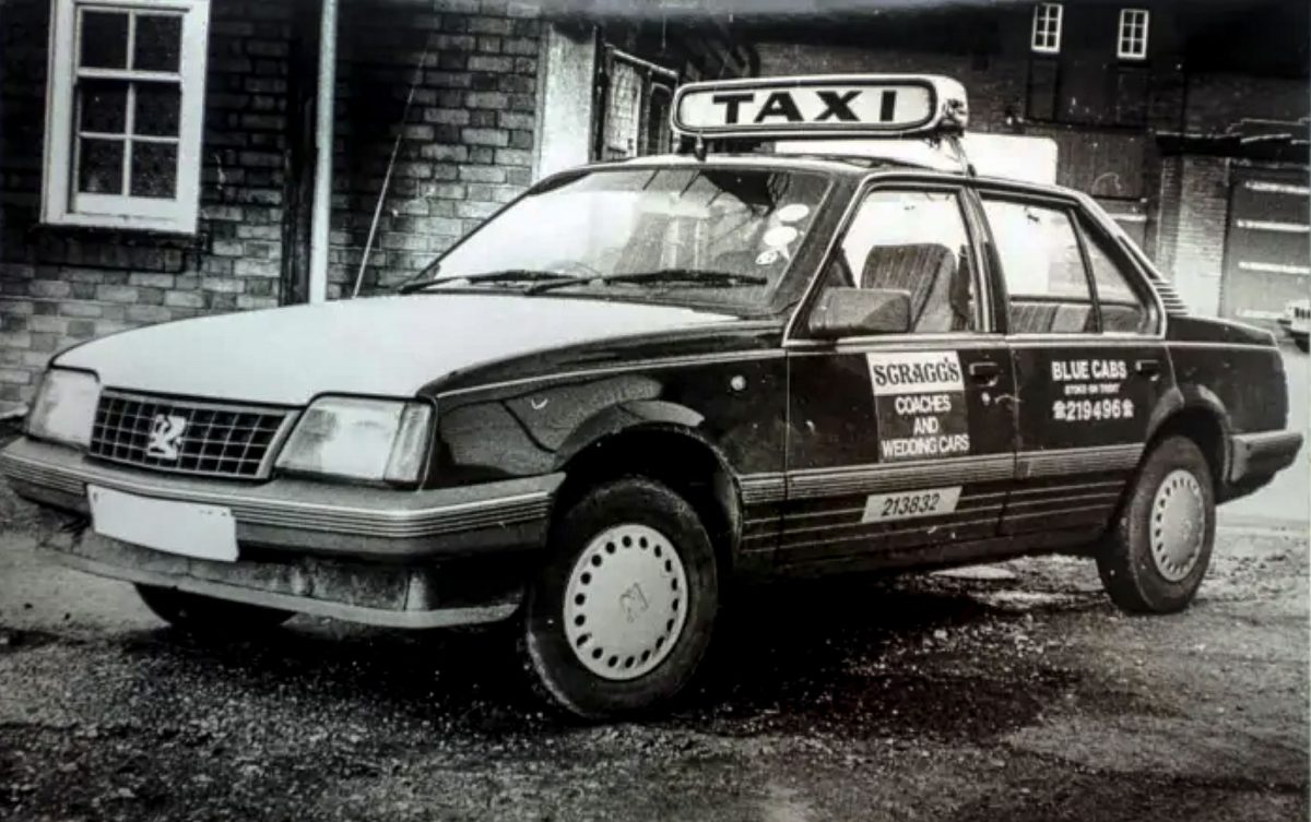 A vintage black and white photograph of a dark Vauxhall Cavalier with a "TAXI" sign on the roof and "Scragg's Coaches" branding on the door.