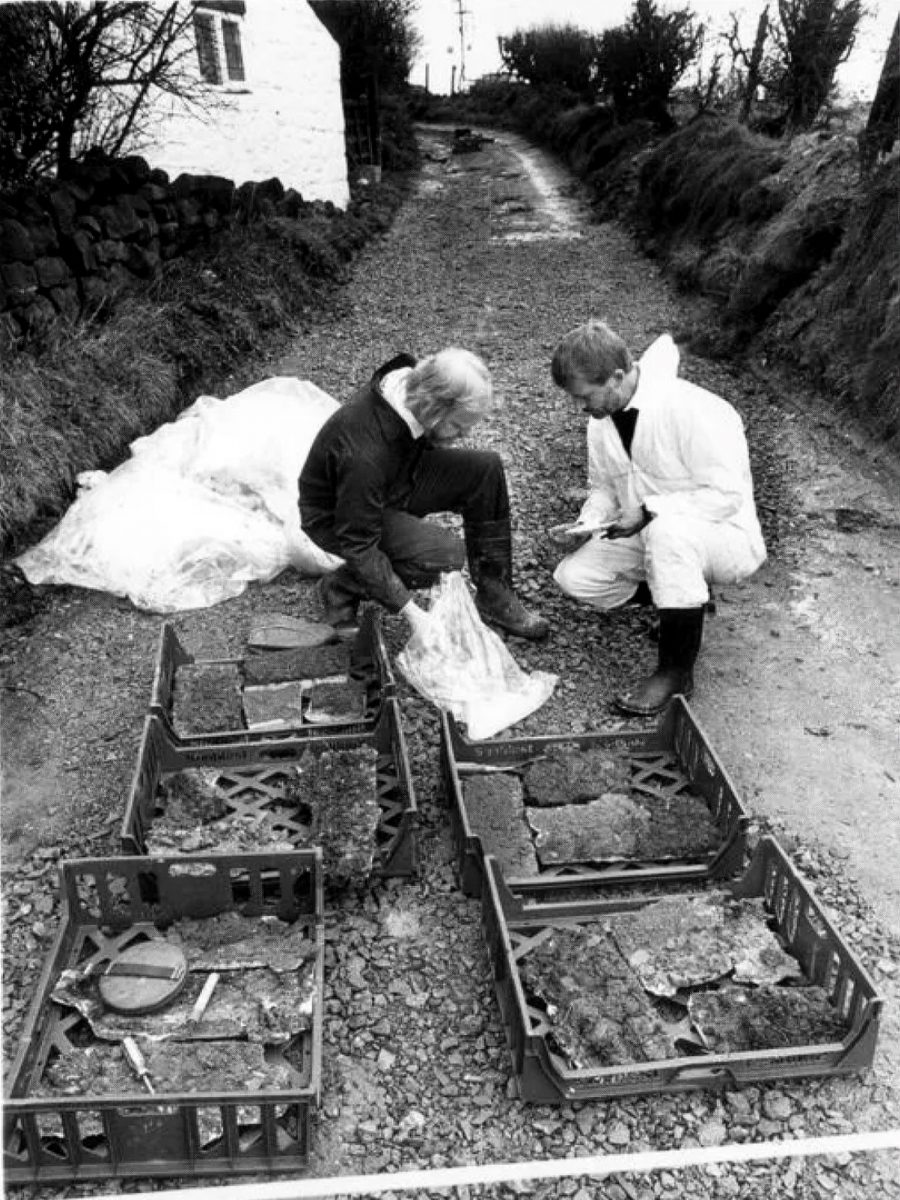 Two men in work gear kneeling on a gravel road next to plastic crates containing samples and evidence bags.