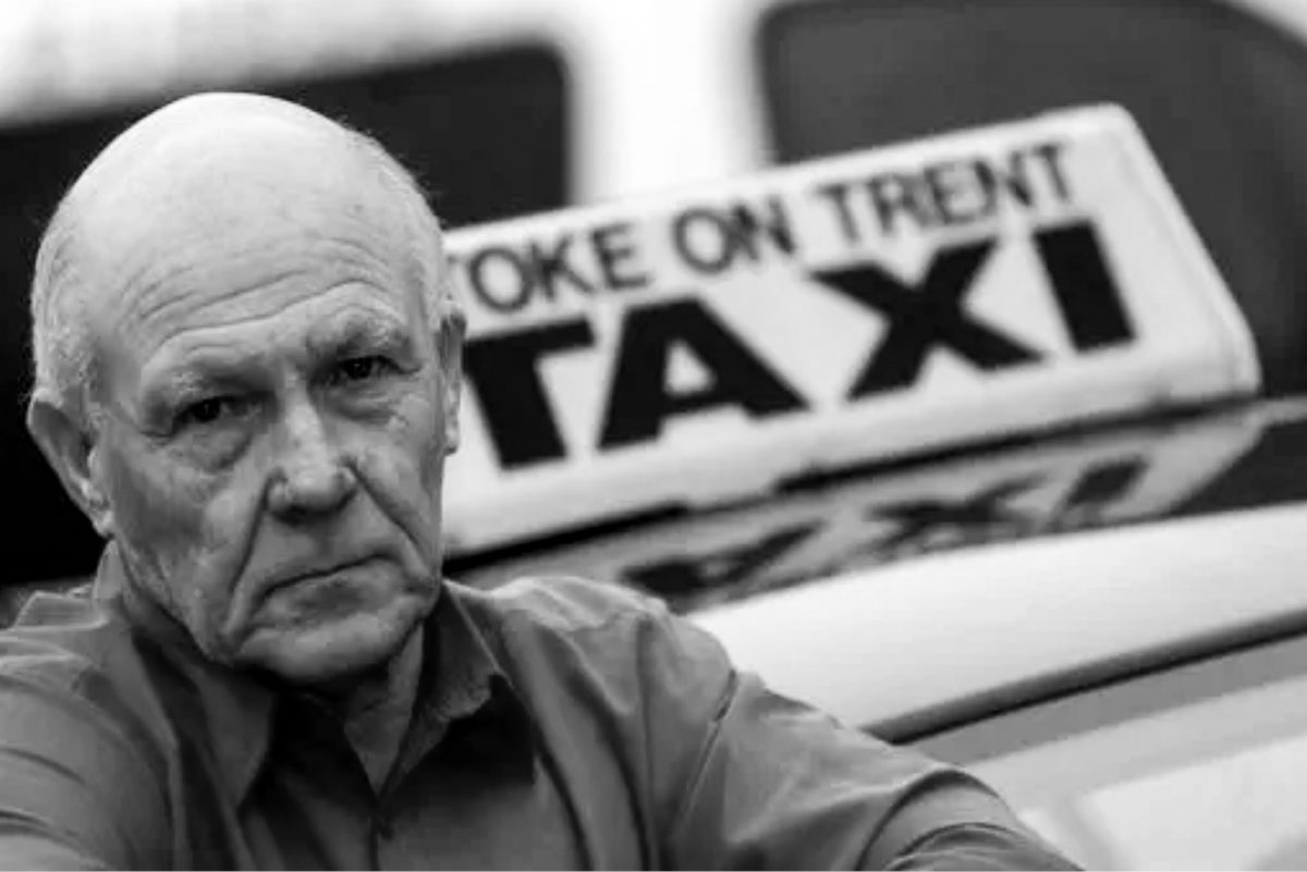A close-up portrait of an older man with a serious expression, with a Stoke-on-Trent taxi sign blurred in the background.