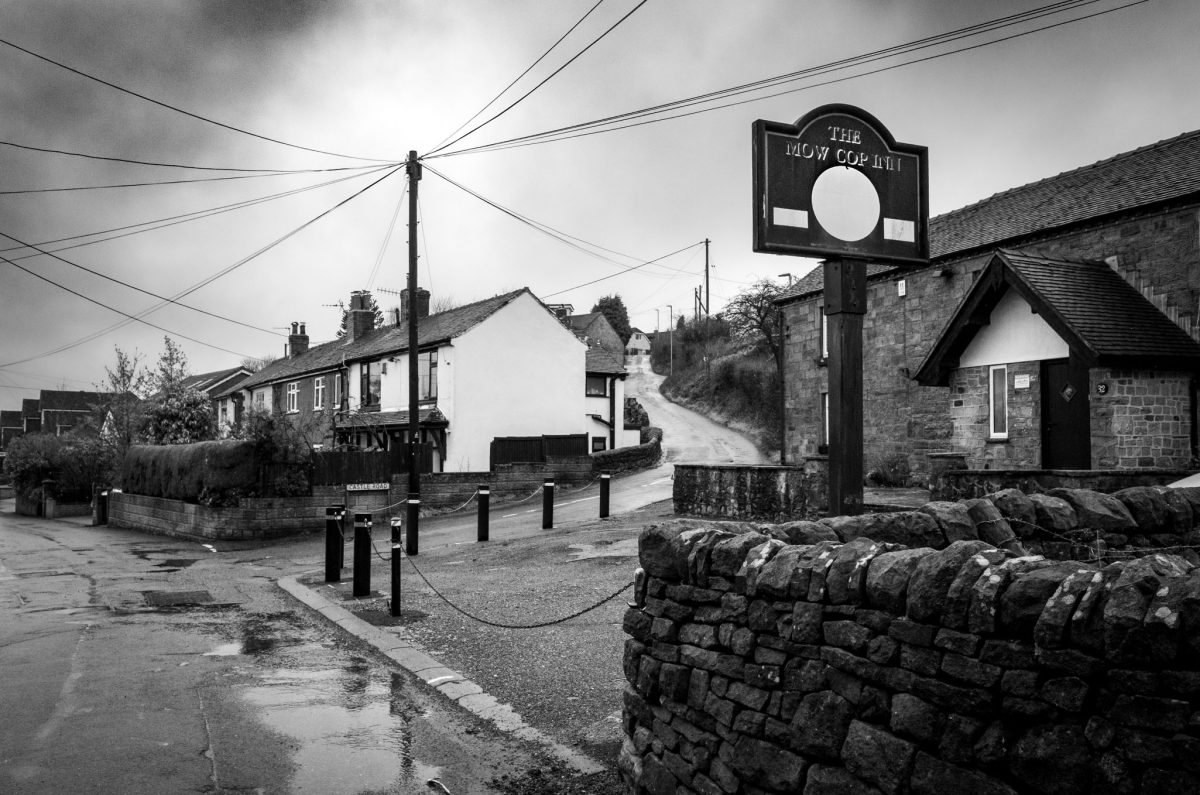 A black and white shot of a stone pub called The Mow Cop Inn with a large sign in the foreground and a road winding up a hill.
