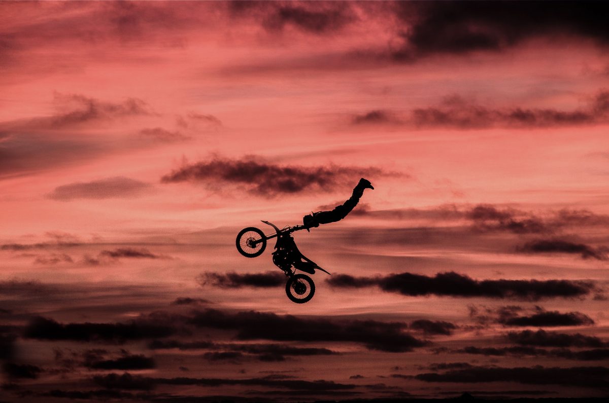 Extreme sports silhouette of a dirt bike stunt rider mid-air, performing a Superman or similar jump trick against a dramatic, fiery red and black sunset sky.