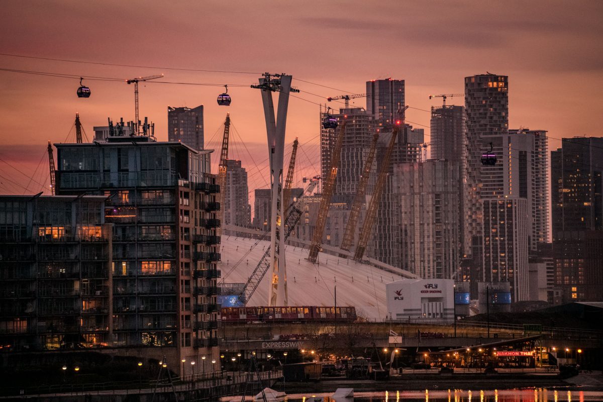 Dramatic sunset over London skyline featuring the illuminated O2 Arena dome, surrounding high-rise buildings, and the Emirates Air Line cable cars.