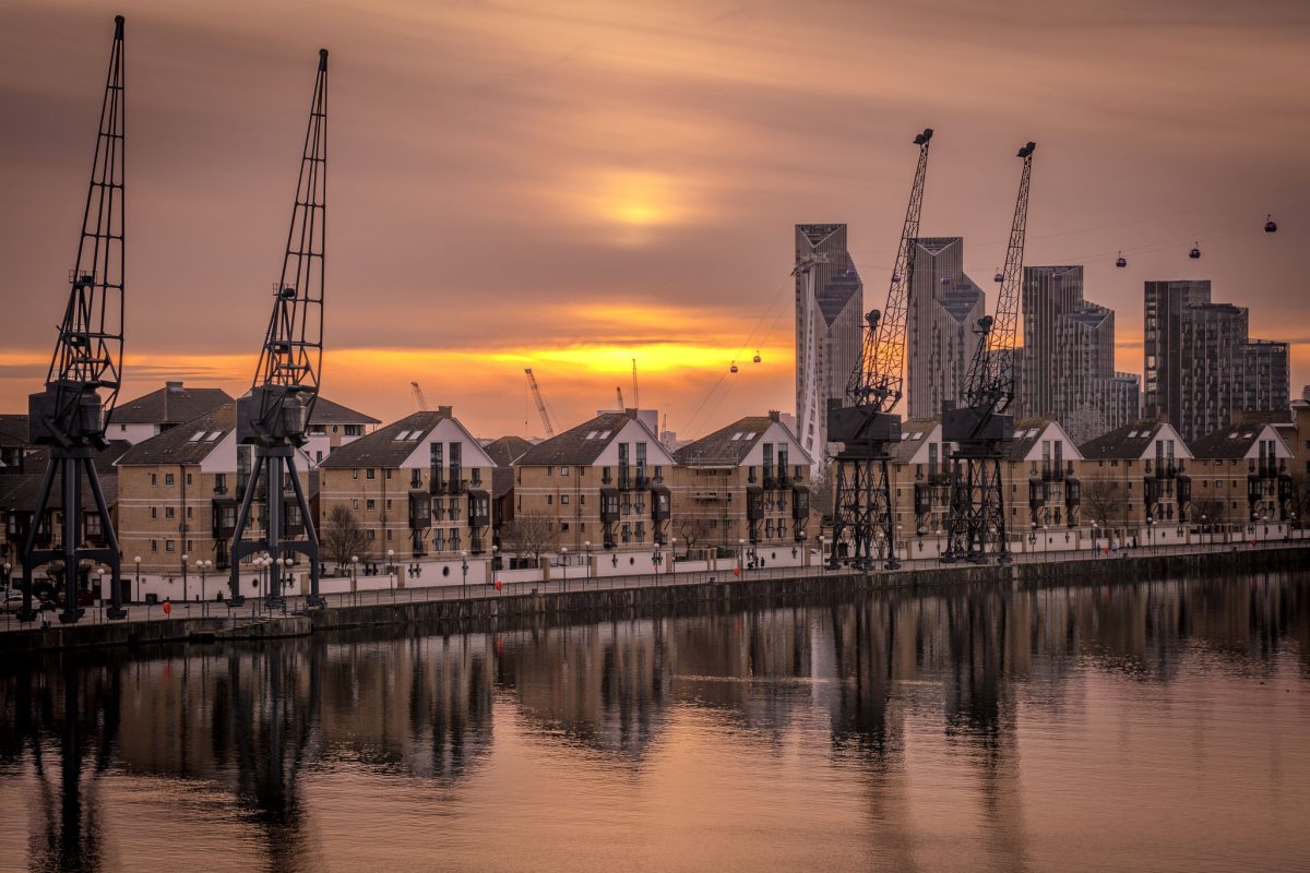 Sunset view of Royal Victoria Dock in London, featuring rows of modern waterside apartments, several historic black dock cranes, and the reflection of the orange sky in the still water, with Canary Wharf skyscrapers and the Emirates Air Line cable cars visible in the background.