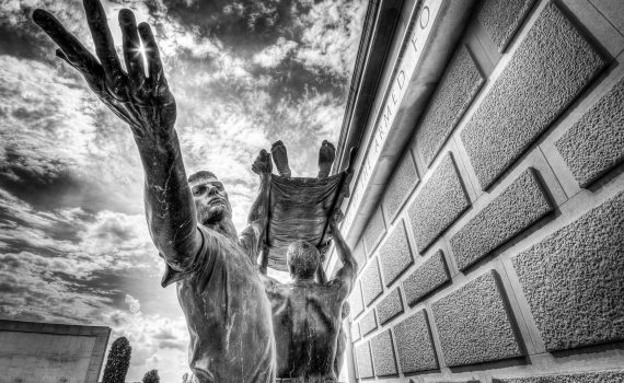 Black and white photo of the Stretcher Bearers Memorial at the National Memorial Arboretum, showing two figures lifting a stretcher beneath dramatic clouds.