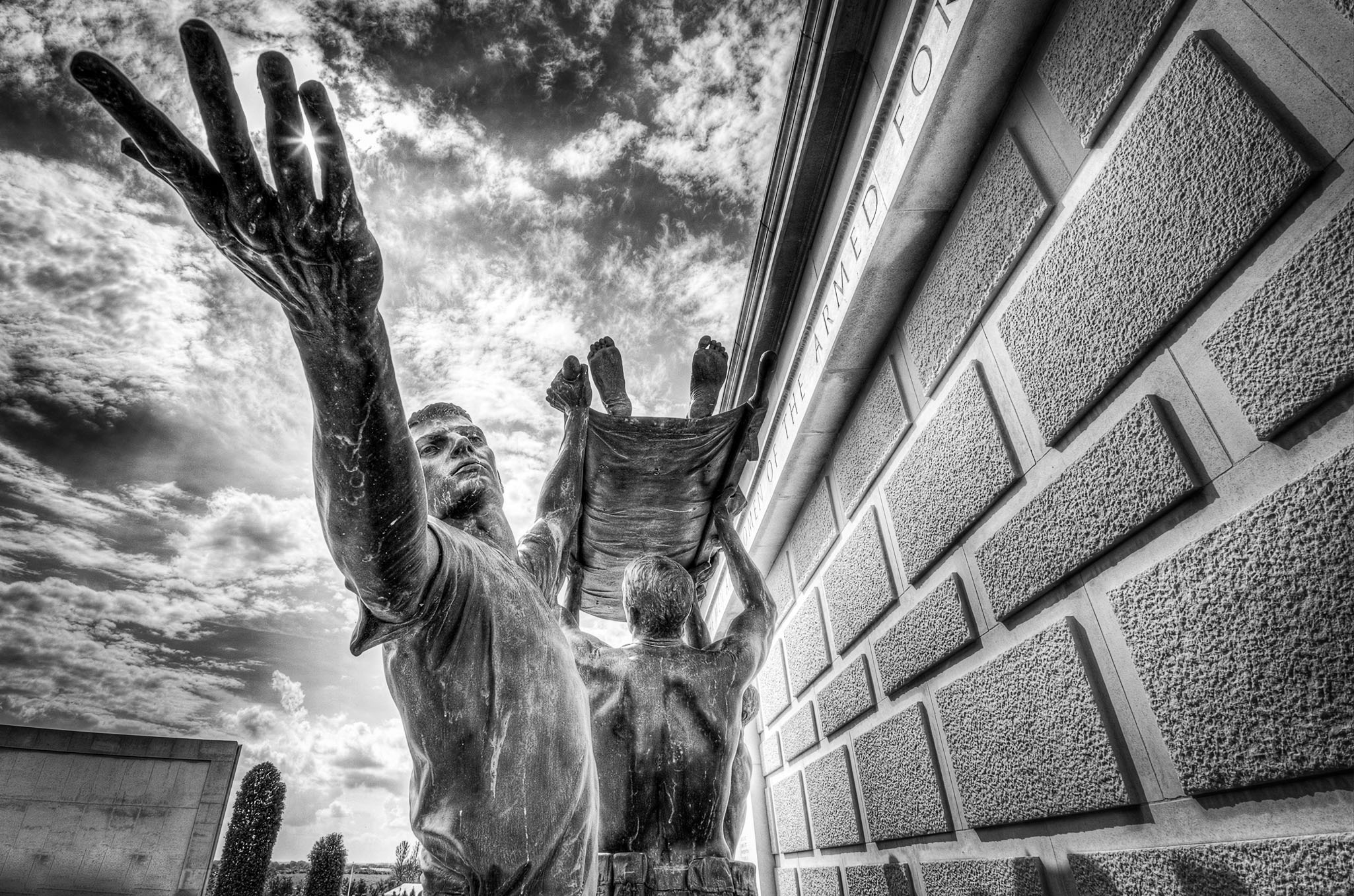 Black and white photo of the Stretcher Bearers Memorial at the National Memorial Arboretum, showing two figures lifting a stretcher beneath dramatic clouds.