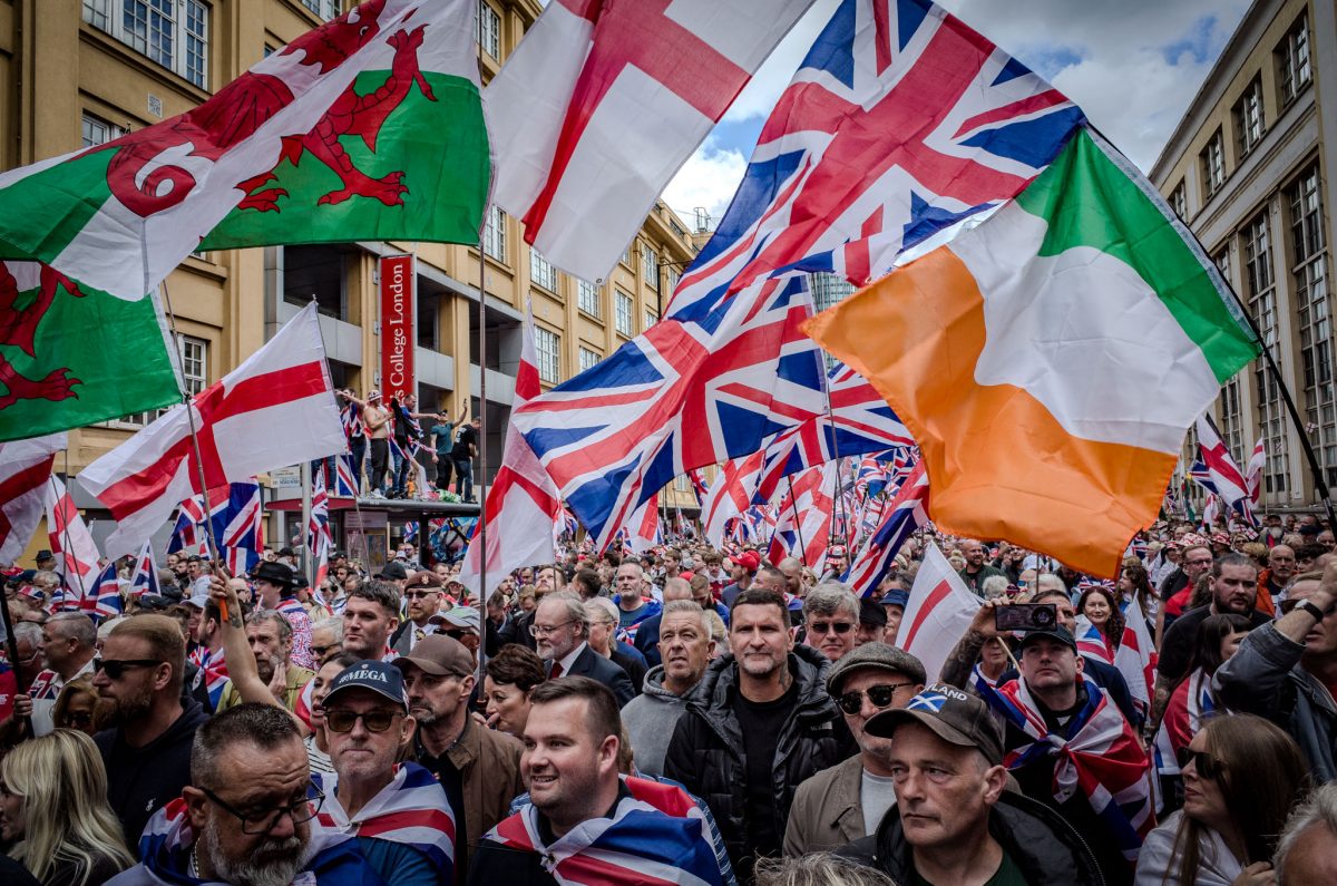 A wide shot capturing a large, crowded political rally in a city street, dominated by people holding high various national flags of the UK, including the Union Jack, the English flag, and the Welsh flag, with buildings visible in the background.