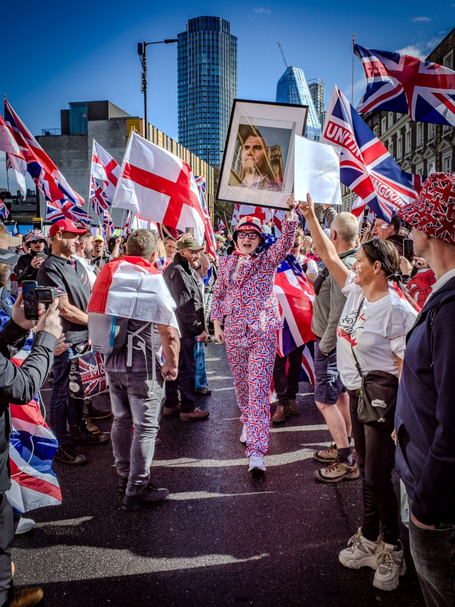 A full-body shot of a woman in a distinctive Union Jack print jumpsuit and sunglasses, holding up a framed photo above her head. She is surrounded by a large crowd holding numerous English and British flags in a sunny city street.