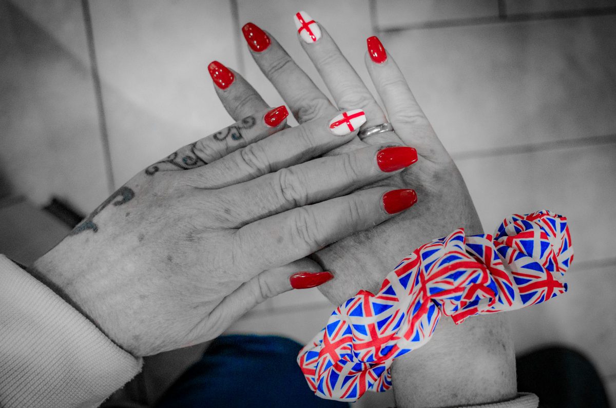 A close-up of two hands. The hands are painted with red nail polish, with one fingernail featuring a white background and a red cross (St. George's Cross). The wrist has a Union Jack patterned fabric scrunchie. The background is desaturated, focusing attention on the colors.
