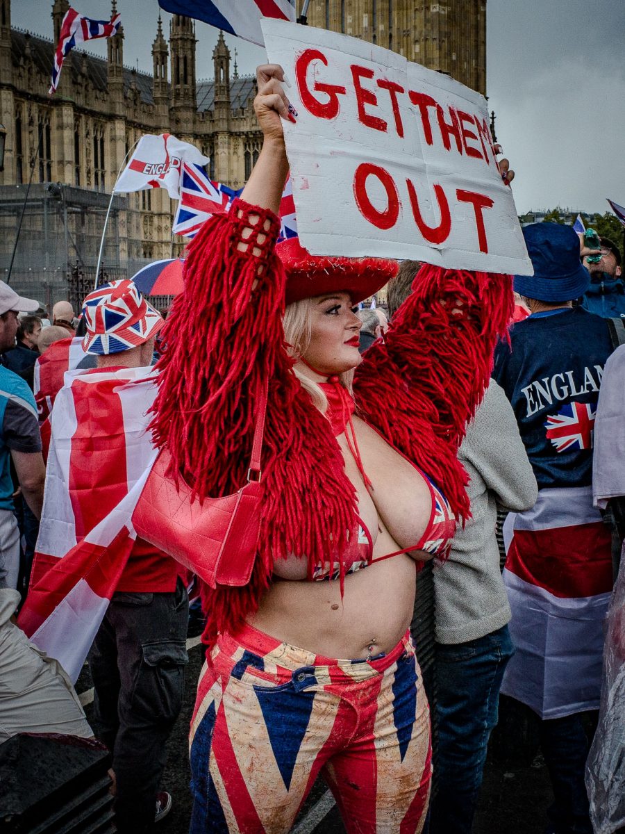 A female demonstrator at a rally holds a sign that reads "GET THEM OUT." She wears a Union Jack-themed outfit, including a bikini top, pants, and a red shaggy coat, with the Houses of Parliament visible in the background.