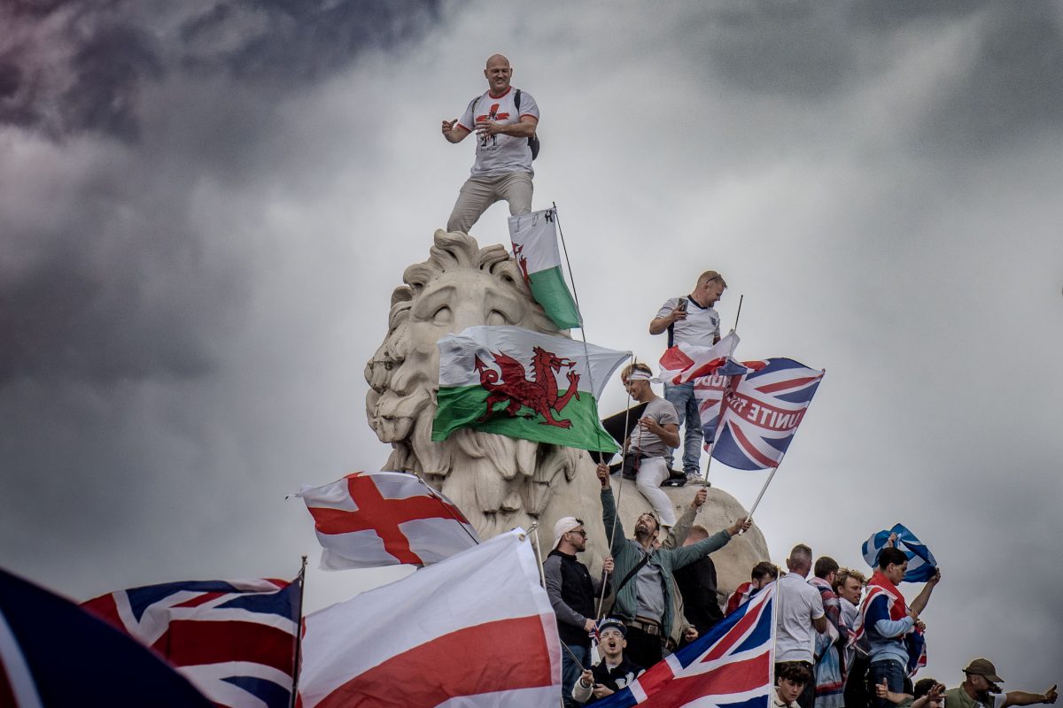 A dramatic, low-angle photograph of several men standing on a large stone lion statue (likely in Trafalgar Square), holding and waving various flags including the Welsh Dragon and the Union Jack, set against a brooding, cloudy sky.