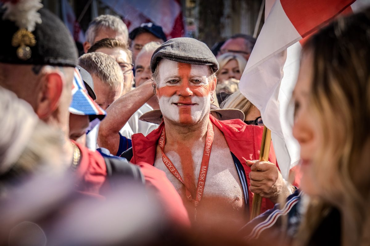 A close-up portrait of an older male demonstrator at a rally. He is smiling directly at the camera with his face painted white and red in the pattern of the English flag, holding a flag pole, and wearing a red vest and a flat cap.