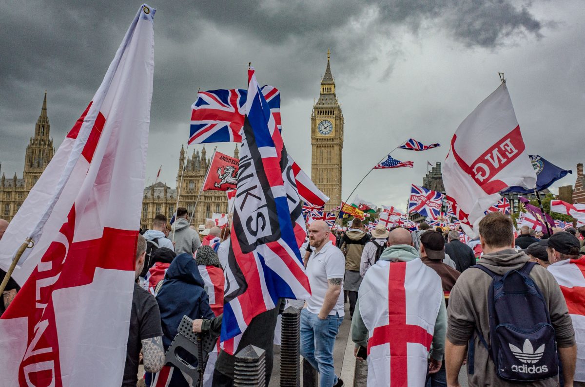 A photograph capturing a large group of people marching in London, with numerous St. George's Cross flags and Union Jacks being carried. The Houses of Parliament and the Elizabeth Tower (Big Ben) are prominent landmarks in the background under an overcast sky.
