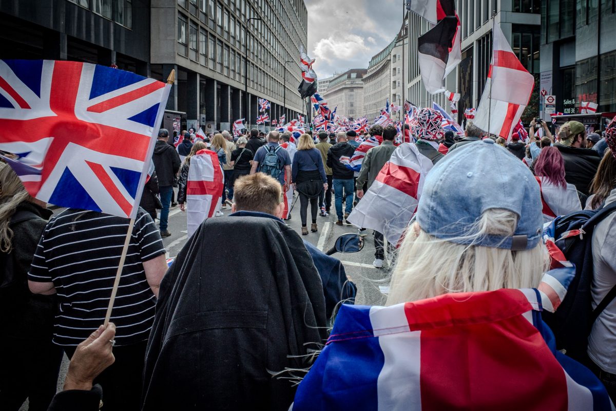 A medium shot of a large crowd marching down a street lined with modern buildings. The foreground is dominated by the backs of participants, many holding English and British flags, including a Union Jack flag on the left and a person wearing a Union Jack cap in the lower right.