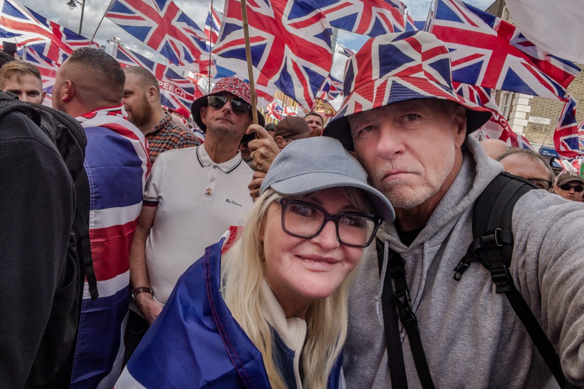 A close-up selfie of a smiling woman and a man at the rally. The man is wearing a Union Jack bucket hat, and the woman is wearing glasses and a light blue baseball cap, draped partially in a Union Jack flag.
