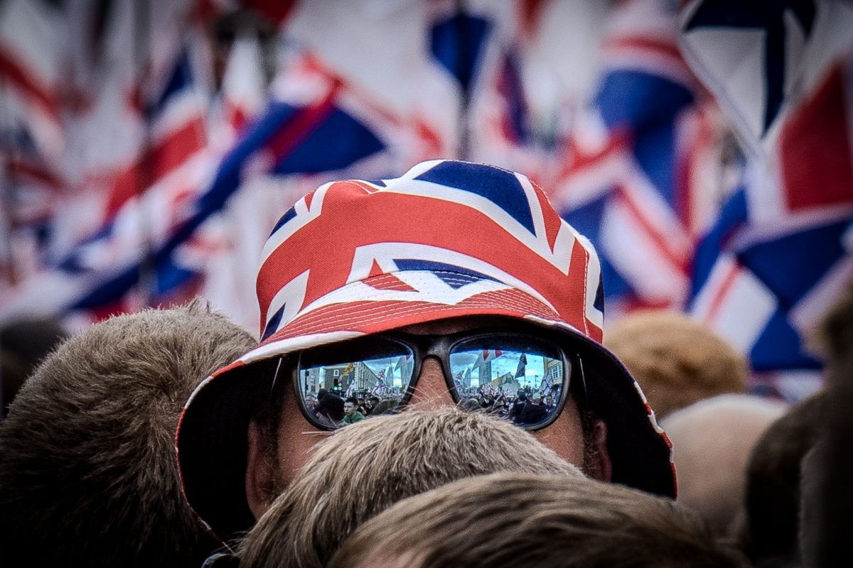 A tight close-up focused on a person's head, obscured by a Union Jack-patterned bucket hat and mirrored sunglasses. The reflection in the lenses clearly shows the crowd and a sea of flags surrounding the individual.