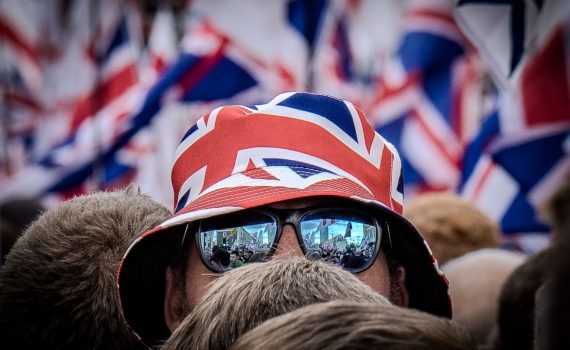 A tight close-up focused on a person's head, obscured by a Union Jack-patterned bucket hat and mirrored sunglasses. The reflection in the lenses clearly shows the crowd and a sea of flags surrounding the individual.