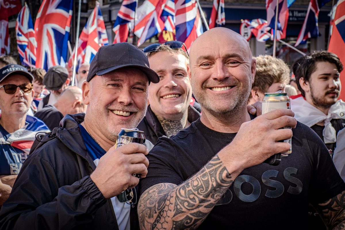 A close-up group portrait of three male demonstrators. The two men in the foreground are smiling and holding cans of drink, with the man on the right featuring a large sleeve tattoo. The background is a dense collection of Union Jack flags.