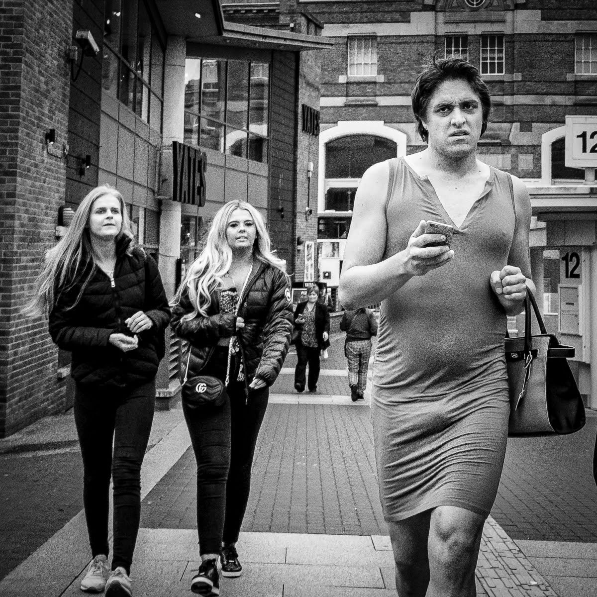 Black and white photo of a person in a tight dress holding a mobile phone and bag, walking on a city sidewalk past two women with long hair.