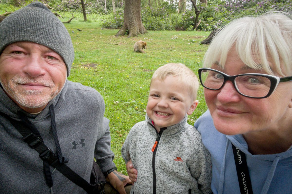 A close-up selfie of a man, a woman in glasses, and a smiling young boy at Trentham Monkey Forest, with a monkey visible in the grassy background.