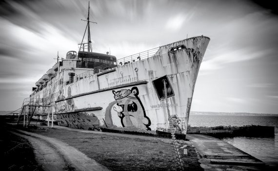 Black and white long-exposure photograph of the rusting, beached TSS Duke of Lancaster ferry, adorned with street art, against a dramatic, streaky sky in Wales.