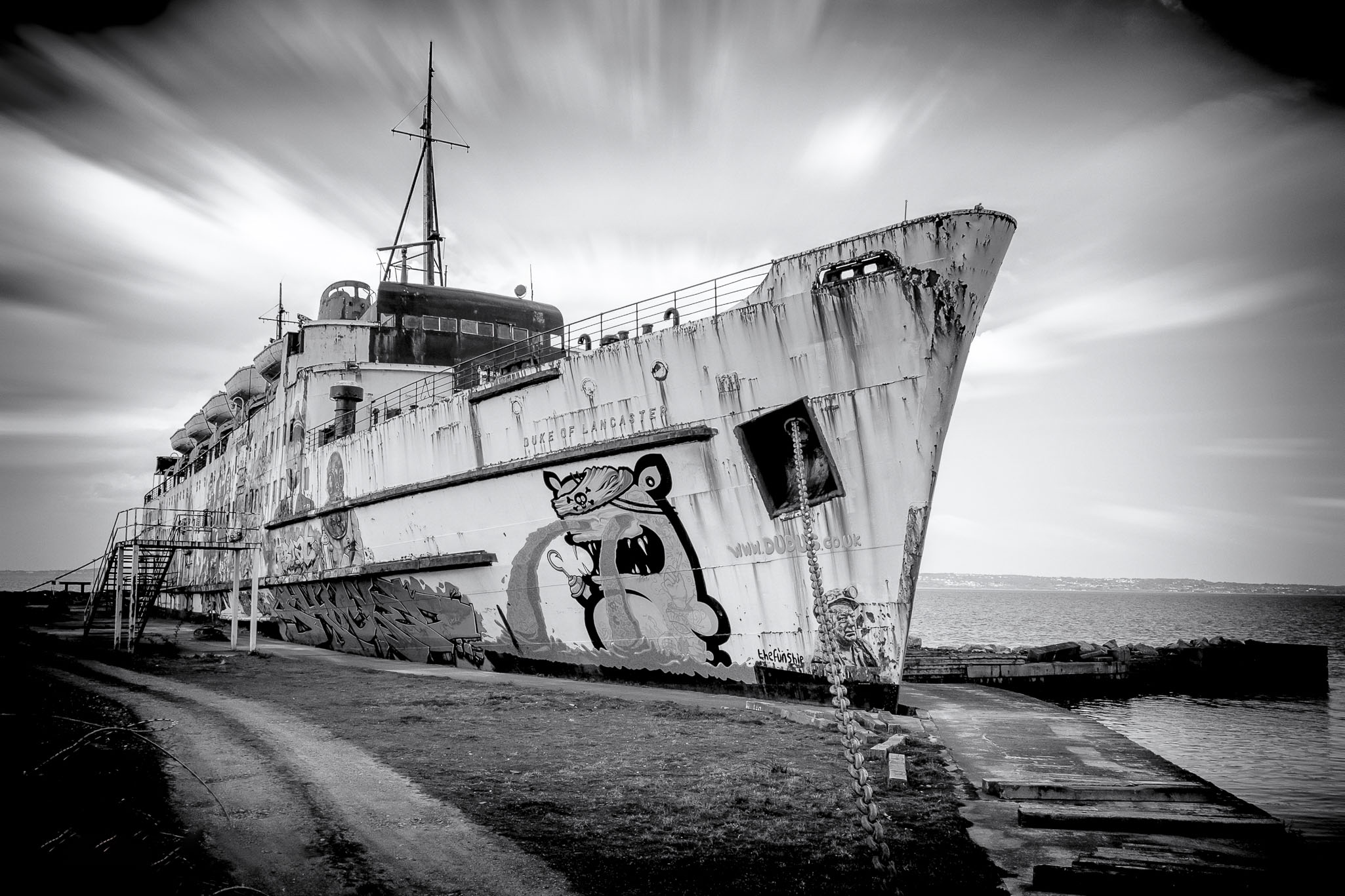 Black and white long-exposure photograph of the rusting, beached TSS Duke of Lancaster ferry, adorned with street art, against a dramatic, streaky sky in Wales.