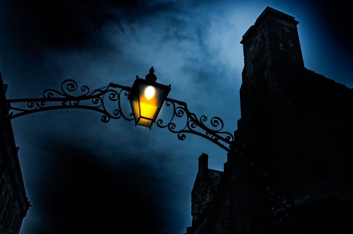 Upward view of a glowing yellow Victorian-style iron lantern and ornate scrollwork against a dark, dramatic blue night sky and silhouetted stone architecture in Edinburgh.