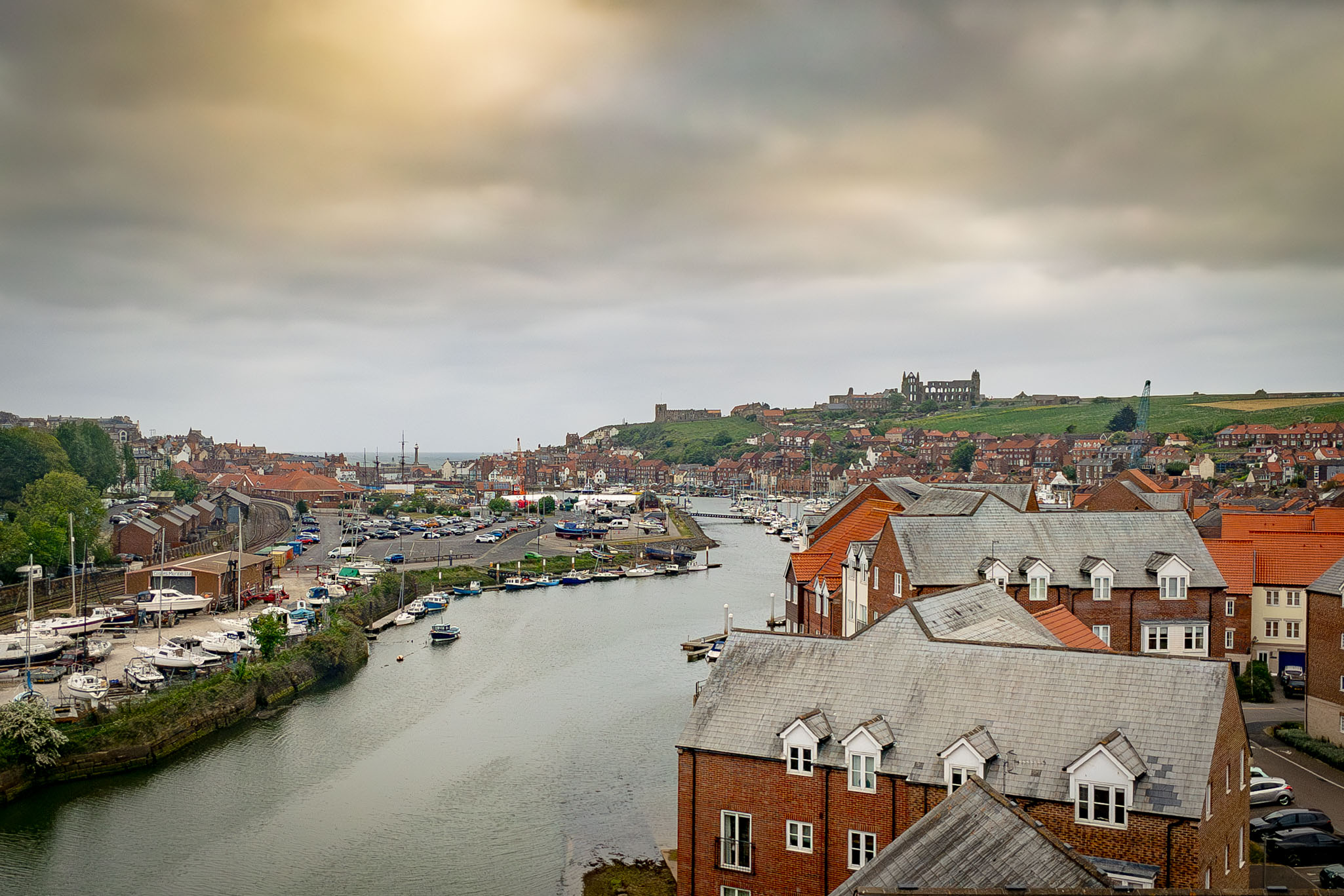 Panoramic landscape view of Whitby harbor, the River Esk, and the town's rooftops with Whitby Abbey visible on the distant hill.