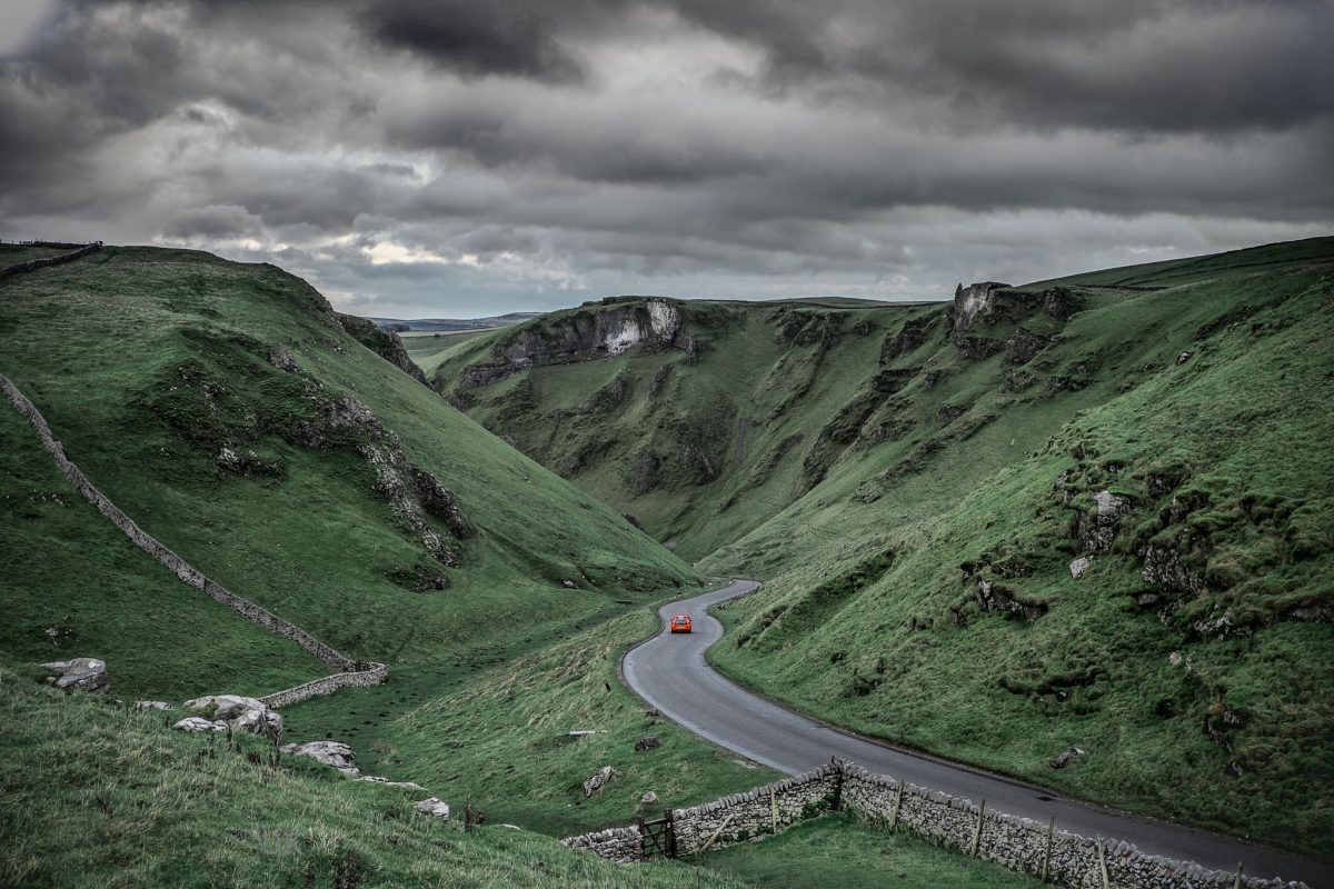 Moody landscape view of Winnats Pass in Derbyshire with a red car driving through the winding gorge under dark storm clouds.