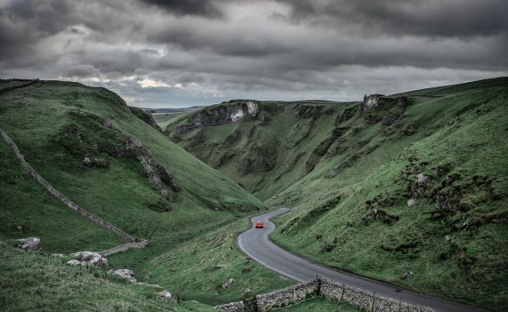 Moody landscape view of Winnats Pass in Derbyshire with a red car driving through the winding gorge under dark storm clouds.
