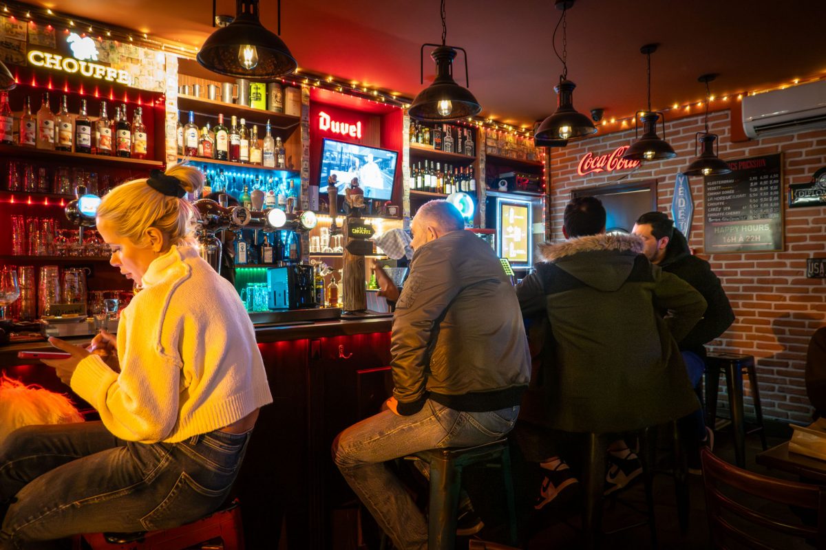 Patrons sitting at the bar inside a dimly lit Parisian pub with red neon accents.